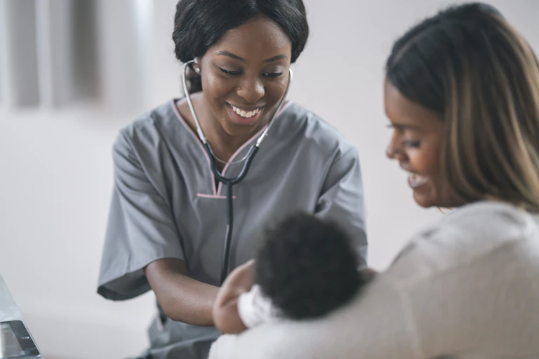 A woman with a stethoscope in her ears smiling at a newborn baby who is being held by another smiling woman lying in a bed