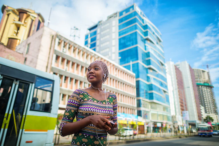 A young, confident-looking woman stands on a prosperous-looking street in Dar es Salaam. She wears white earbuds and a multicolored top; behind her is a gleaming glass building and as bus.