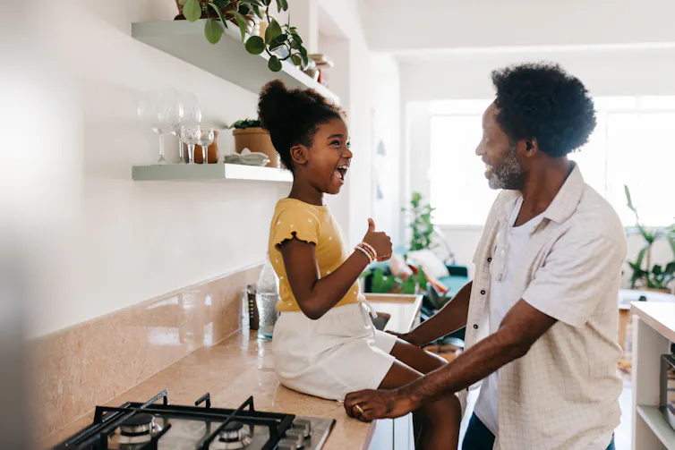 Little Black girl sitting on countertop in kitchen talking to father
