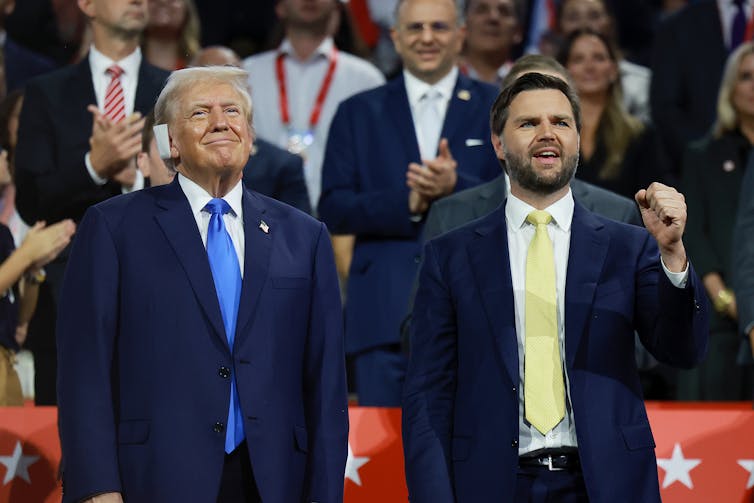 Two white men dressed in business suits stand next to each other on a stage in front of cheering spectators.