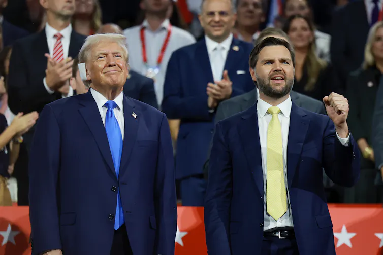 Two white men dressed in business suits stand next to each other on a stage in front of cheering spectators.