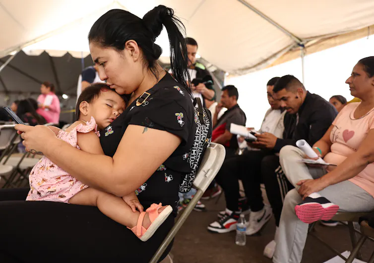 A woman using a cell phone holds a sleeping infant on her lap