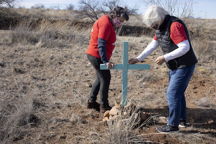 Las aplicaciones, los servicios 911 y los teléfonos móviles no compensan las consecuencias mortales de las políticas fronterizas restrictivas 1 Dos mujeres que llevan naranja están instalando la cruz azul en la tierra arenosa y guisada