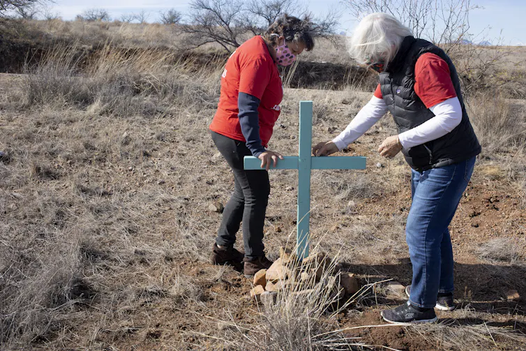 Two women wearing orange install a blue cross into the sandy, scrubby ground