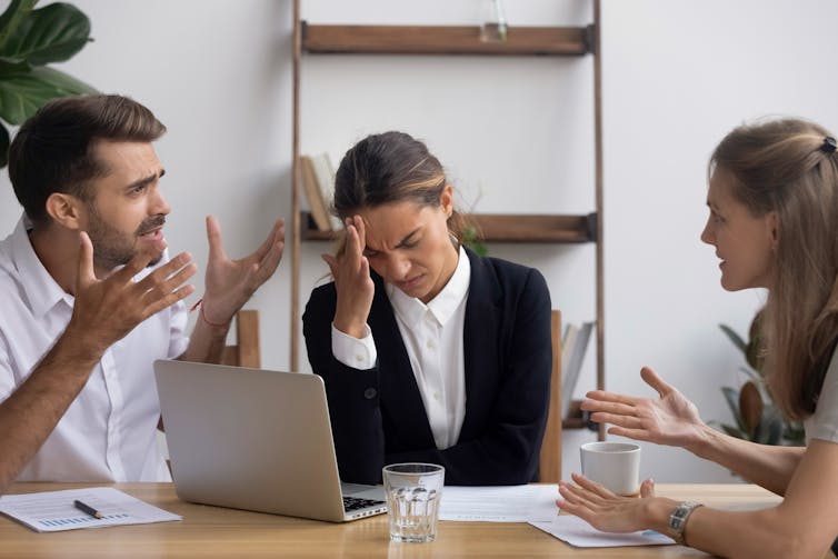 A man and a woman arguing at work while a third woman holds her head in her hands.
