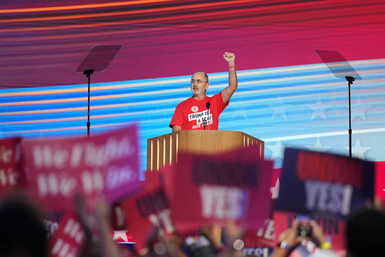 Man wearing red t-shirt holds fist in air from a lectern while a cheering crowd waves red and blue signs.