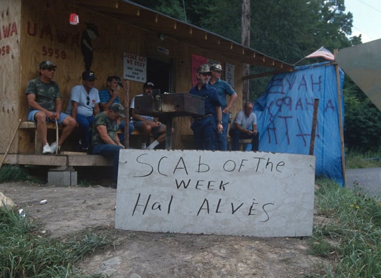 Group of miners wearing baseball hats sit and stand in front of a small building. They've made a sign that reads 'Scab of the week: Hal Alves.'