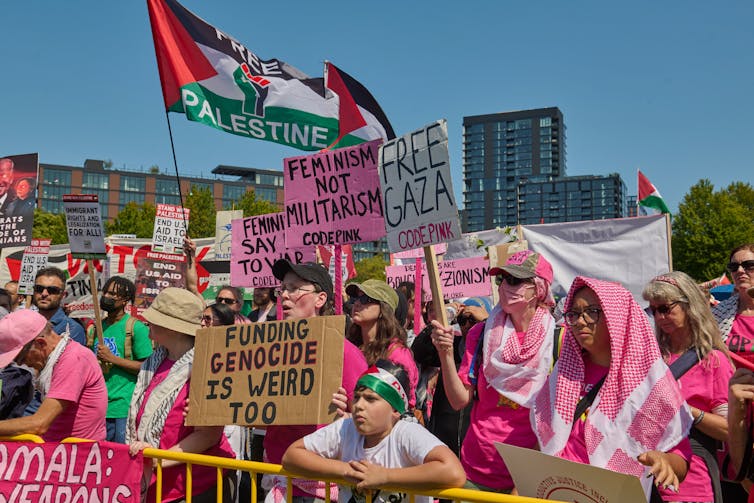 A group of people stand close together and wear mostly pink, with pink and white checkered scarves and hold signs and wave a flag. One sign says 'Free Gaza' and another says 'Funding genocide is weird, too.'