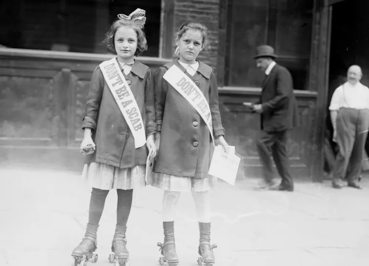 Two young girls on roller skates distribute leaflets while wearing sashes that read 'Don't Be A Scab.'