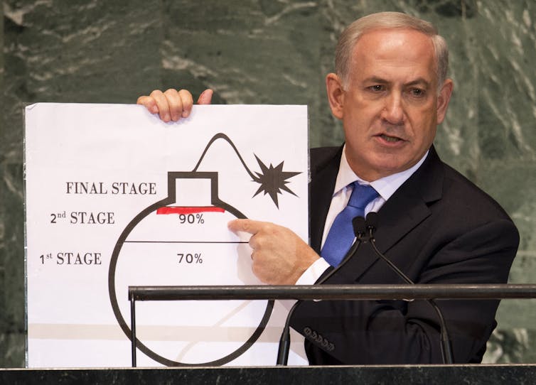 A man wears a suit and points to a picture that shows a bomb that says 'final stage' with a red line near it.