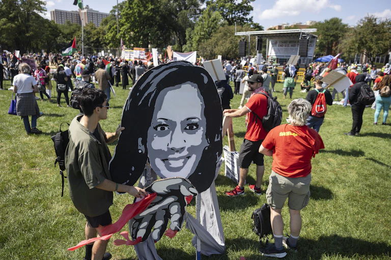 A person holds up a black and white large cutout of a woman's face, while other people stand nearby with signs on a grassy lawn.