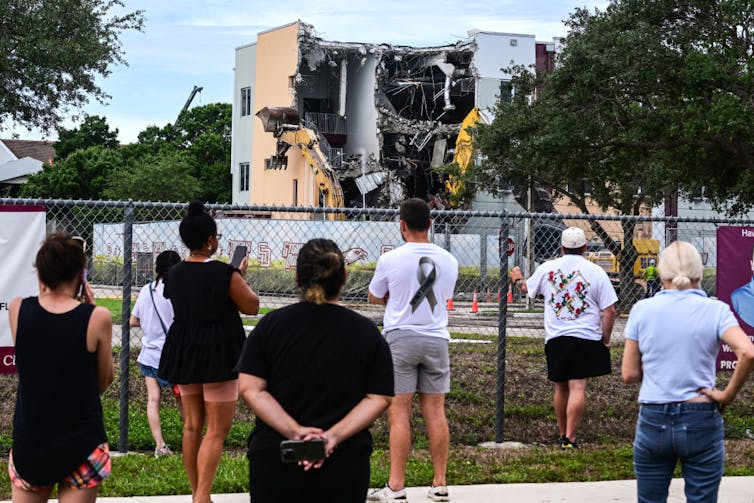 People behind a chain link fence watch as a demolition truck destroys a building.