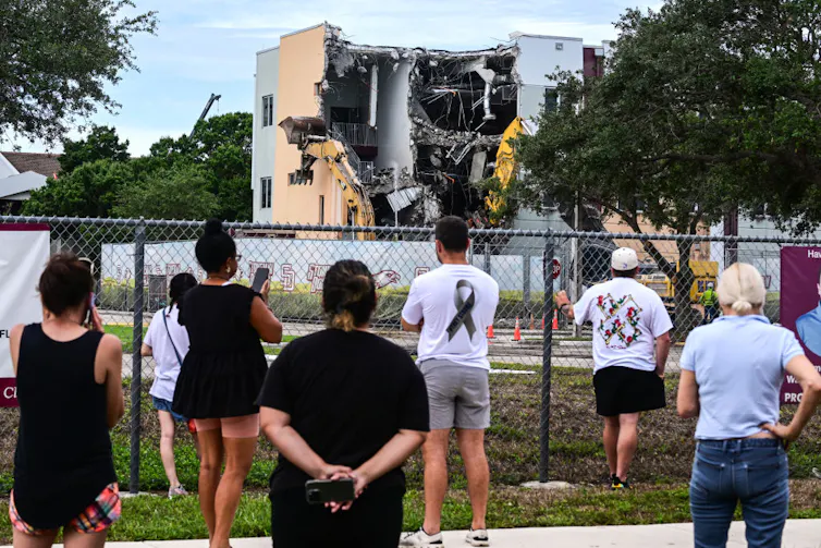 People behind a chain link fence watch as a demolition truck destroys a building.