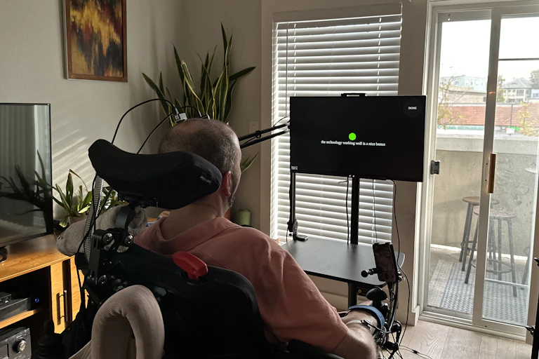 A man in a wheelchair with two wires attached to his head looks at text on a computer screen