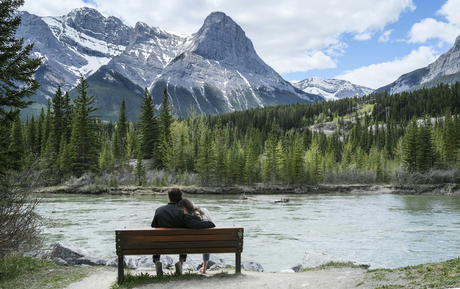 A couple sit on a park bench overlooking a river.