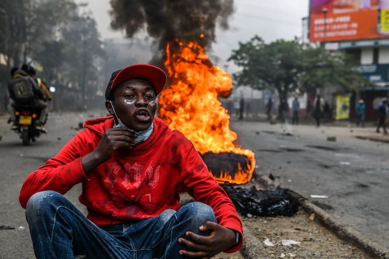 A protestor in a red hat and hoodie sits in front of a fire.