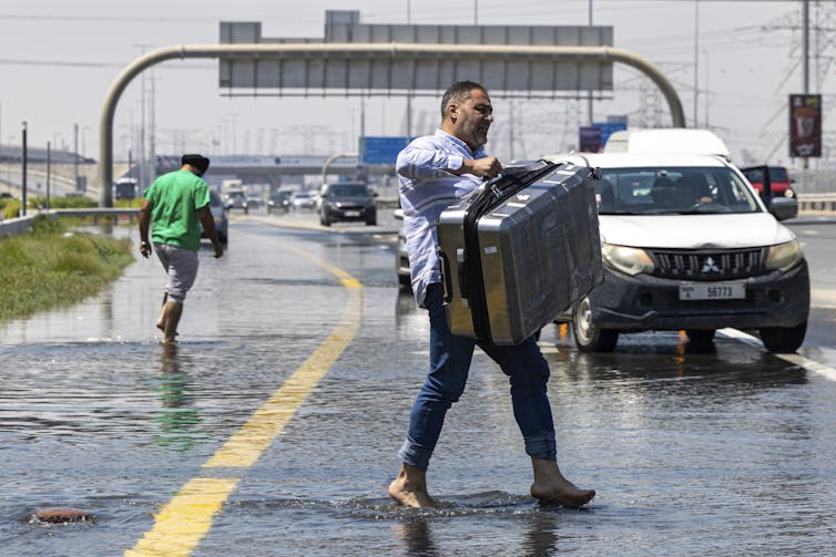 A man walks barefoot across a flooded road carrying a suitcase