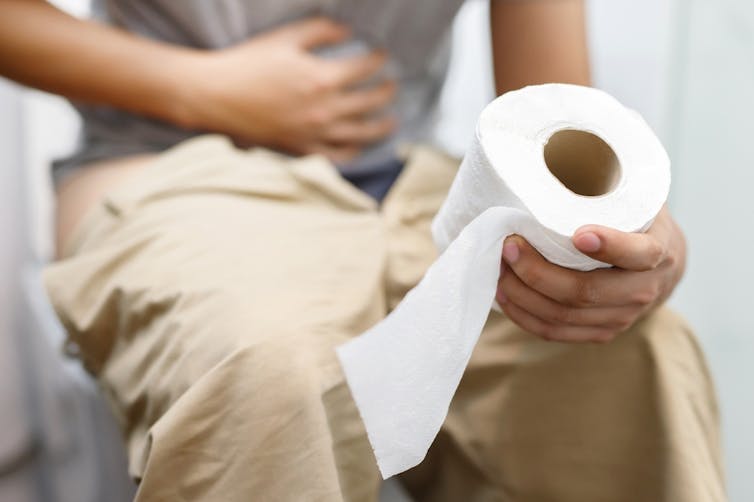 Man sitting on toilet, clutching tummy with one hand, holding toilet roll in other