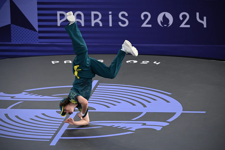 A woman in a green track suit and white sneakers performs an athletic stunt.