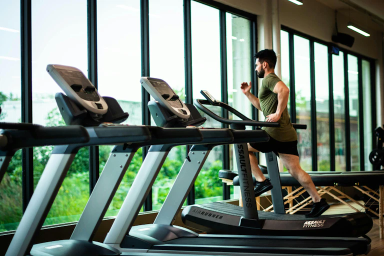 A man in a gym running on a treadmill by a window.