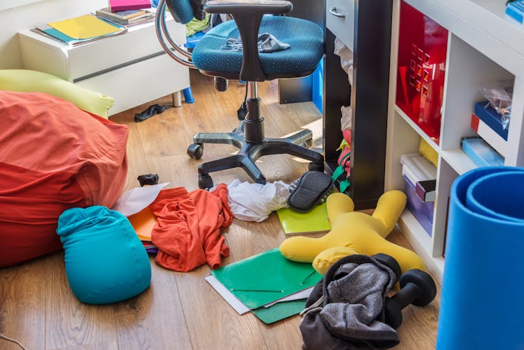 Clothes, papers and books on the floor, near a desk chair and shelves.