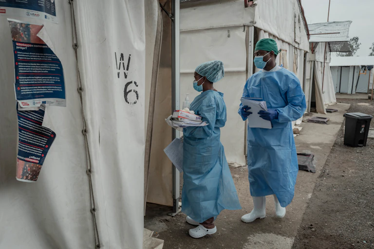 A man and woman wearing medical scrubs and full protective gears