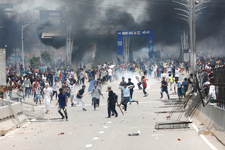 Protestors clash in the middle of a road in Bangladesh as black smoke billows behind them.