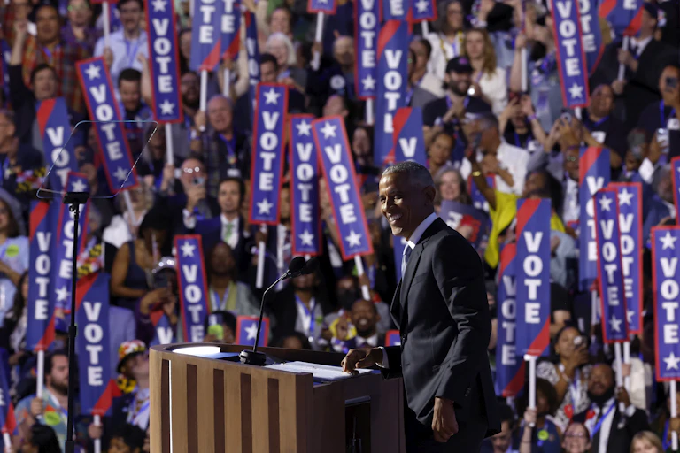A Black man in a suit stands on a stage in front of a crowd waving signs that say 'vote.'