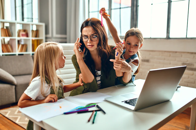 A business woman and a mother are trying to work on a laptop when her little daughters are playing