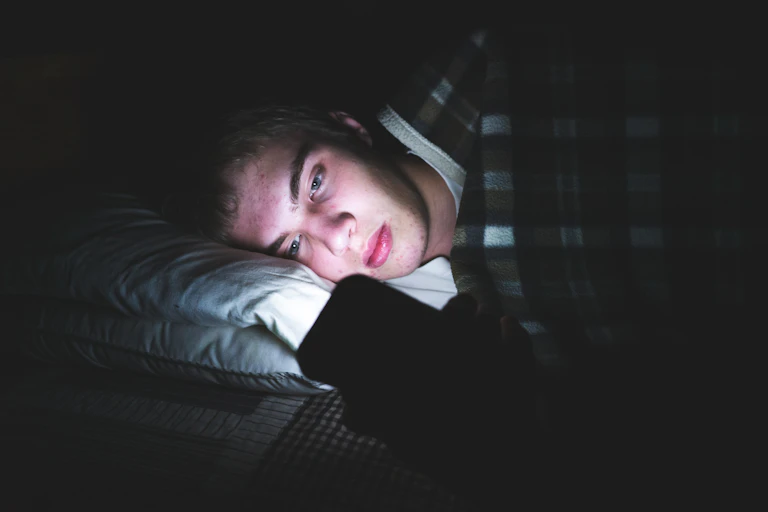 A young man browses the internet on his phone while in bed.