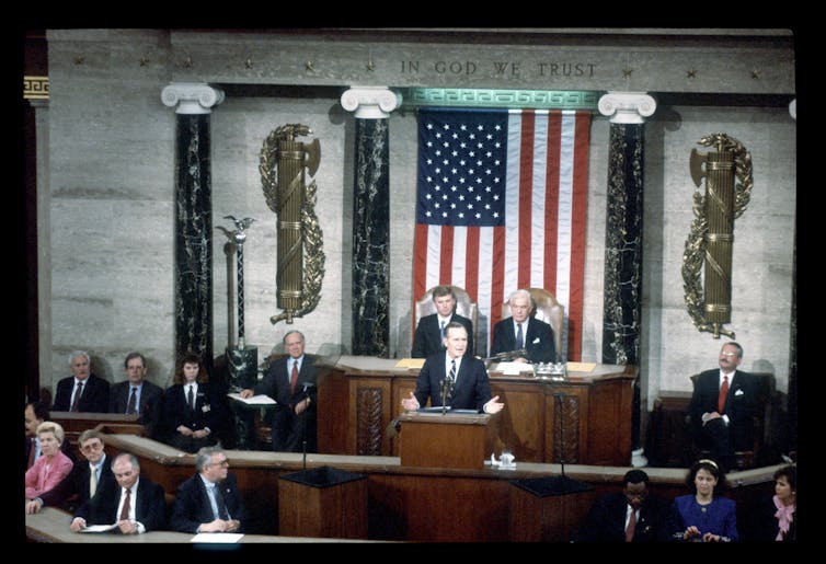 A man stands at a podium behind a large American flag and people seated nearby him.