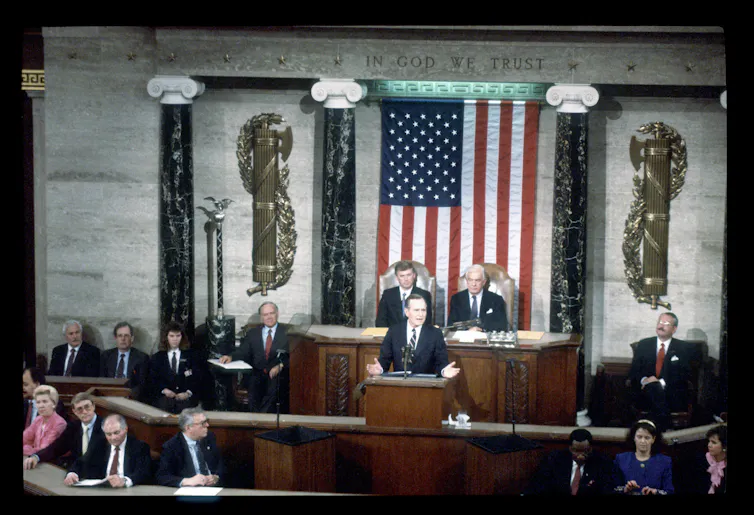 A man stands at a podium behind a large American flag and people seated nearby him.