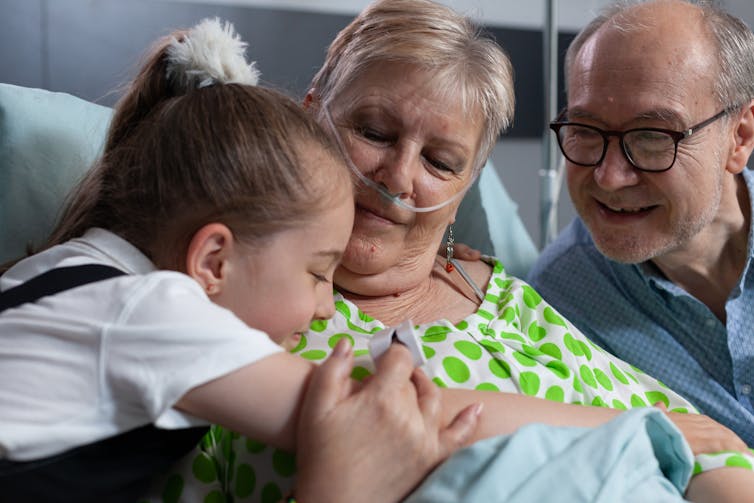 A girl in school uniform hugs an older woman in hospital, while an older man in glasses looks on.