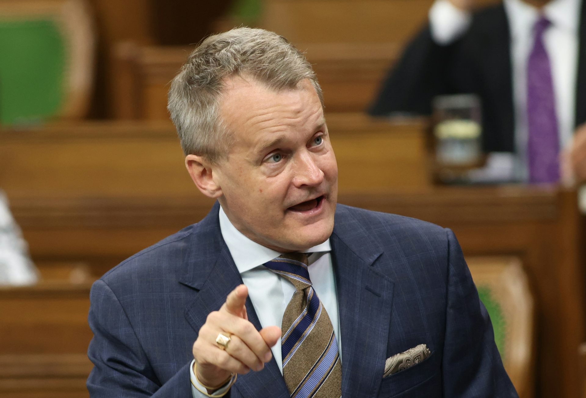 A middle-aged white man with blonde hair points his index finger while speaking to someone off-camera