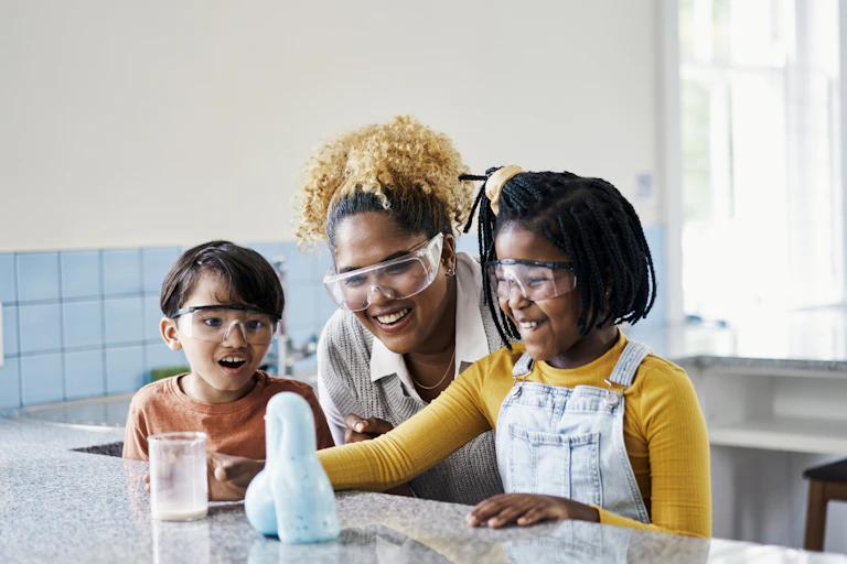 A woman and two young children, all wearing protective goggles, doing a science experiment in a laboratory