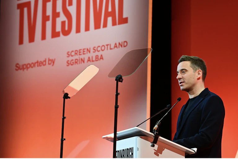 A young man in a black suit delivering a speech on stage with a red background.
