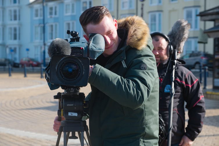 A young guy behind a camera filming a scene in a large square.
