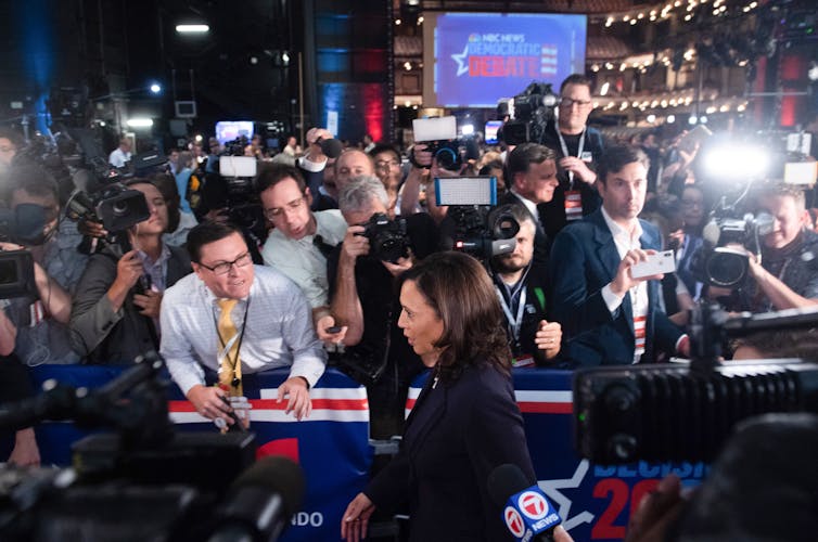 A woman walks past a large group of people holding cameras and microphones.
