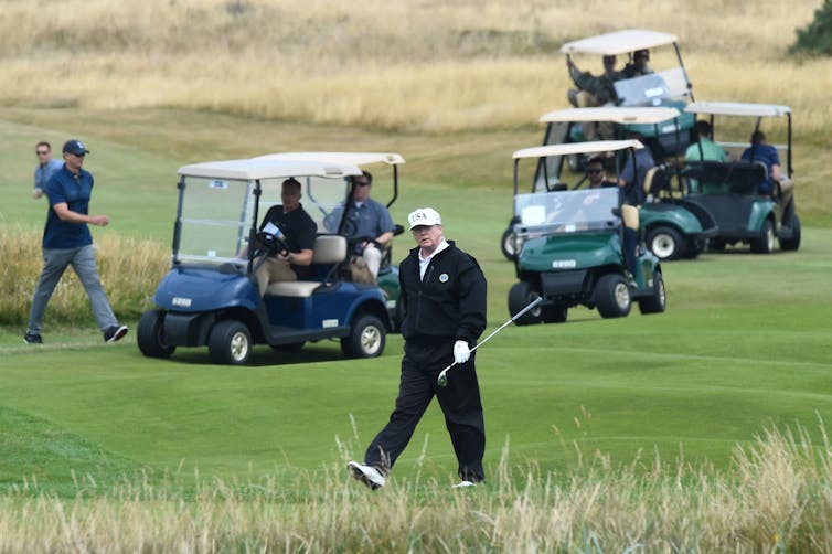 A man wears a black track suit and a white hat and holds a golf club as he walks on grass, surrounded by people who are in golf carts.