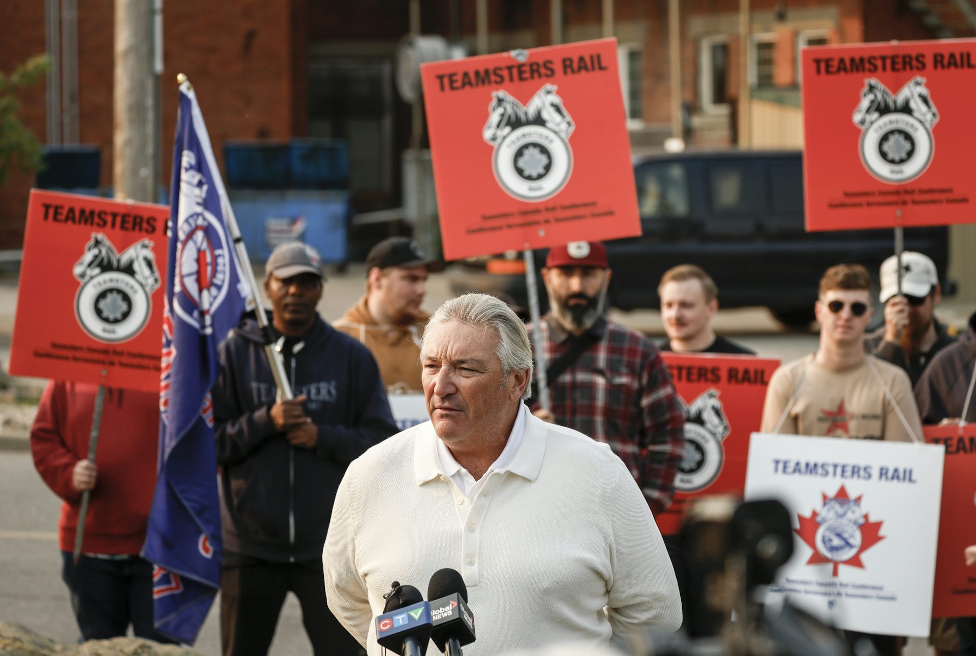 A middle-aged white man in a white shirt speaks to members of the press who are off-camera in front of a crowd of people holding holding 'Teamsters Rail' signs
