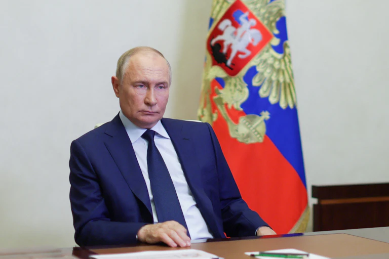 A man in a blue suit sits looking glum in front of a flag.