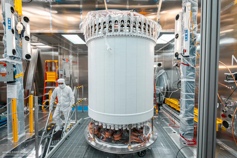 Photo of a person wearing a full-body white suit in a lab next to a large white cylinder.