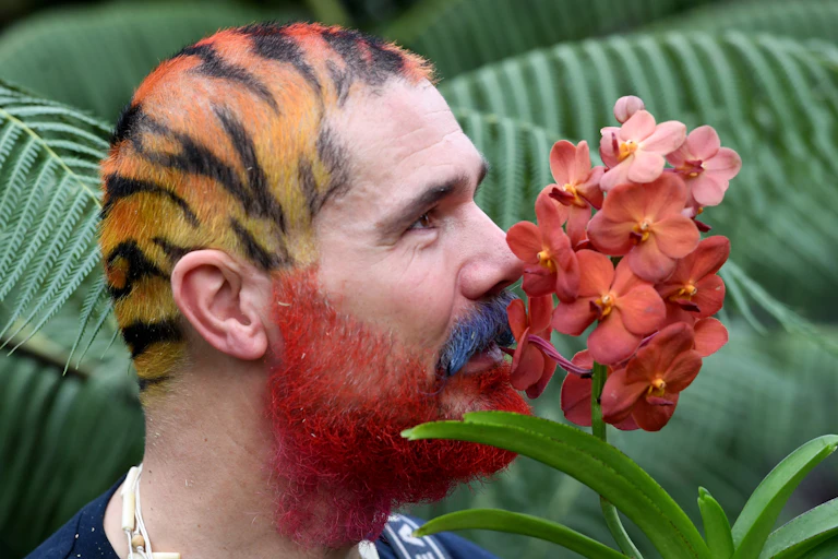 A man with colourful hair and a red bear inhales the scent of an orchid.