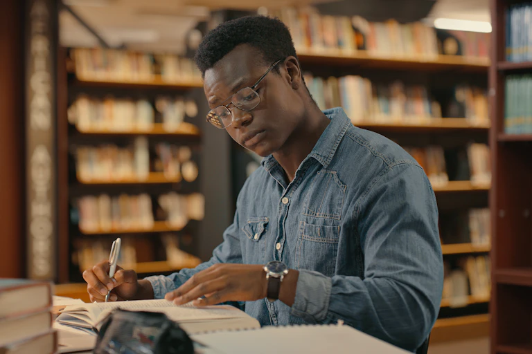 A studious looking young man in a collared shirt working in a library, making notes