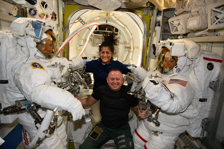 Two smiling people in black shirts next to two people in large white space suits on the ISS.