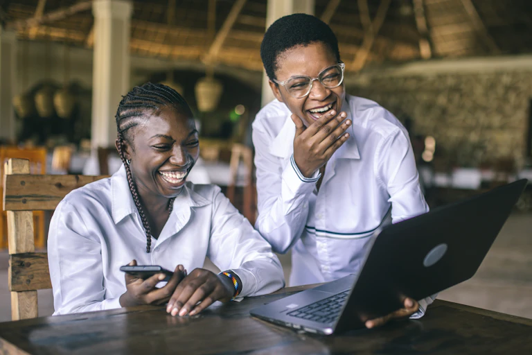 Two young people laugh while looking at a laptop in what appears to be a restaurant setting
