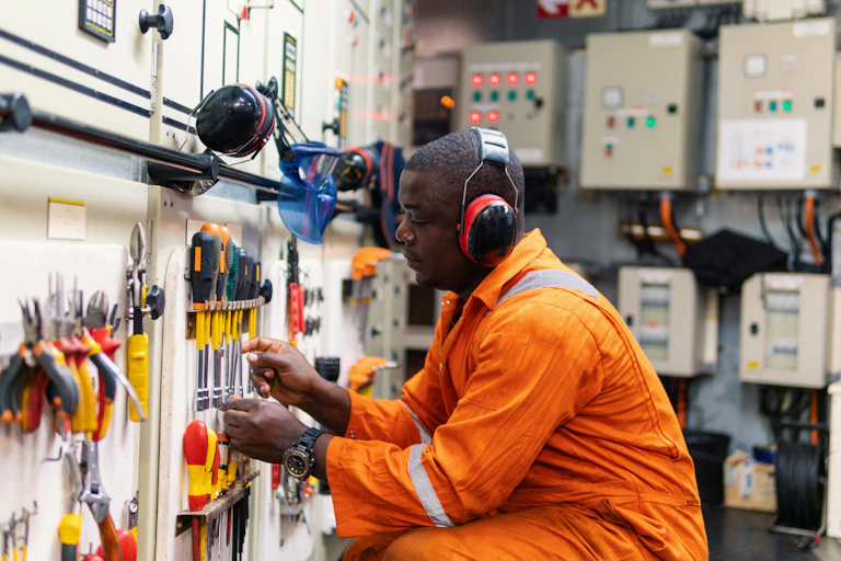 A man in orange overalls, wearing large headphones, works with a variety of tools neatly arranged on a white wall
