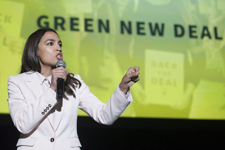 A woman in a white jacket with long dark hair speaks into a microphone with a banner behind her that reads Green New Deal.