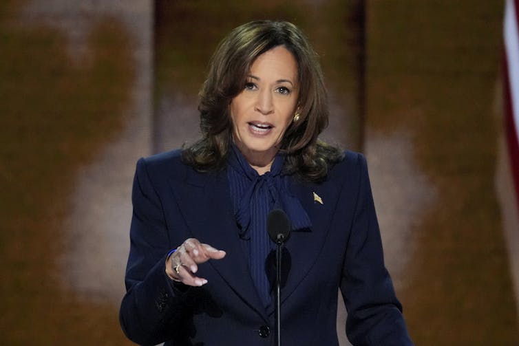 A dark-haired woman in a navy suit gestures as she speaks on a stage.