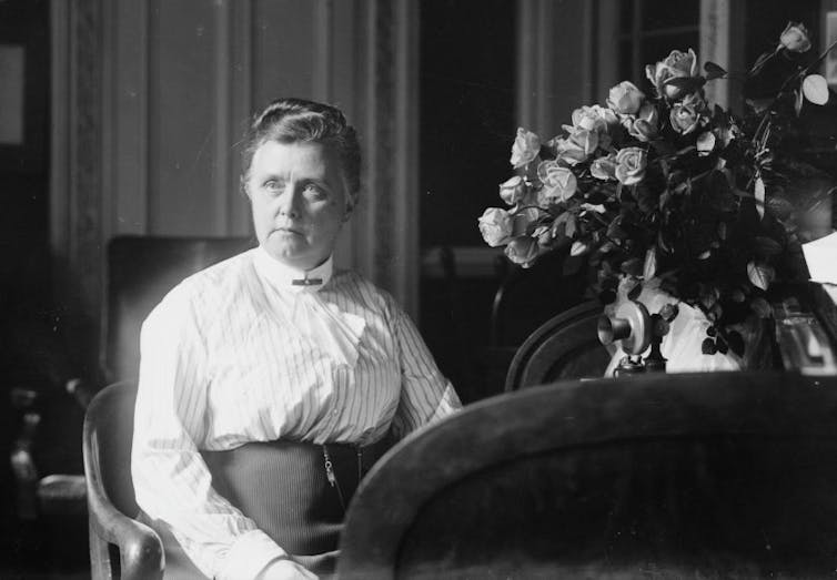 Woman in white blouse seated in chair posing for a portrait next to a bouquet of flowers.